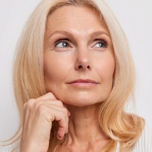 Thoughtful blonde middle aged woman ponders on something keeps hand near face has healthy skin minimal makeup makes choice wears white blouse poses indoor blank copy space for your promotion